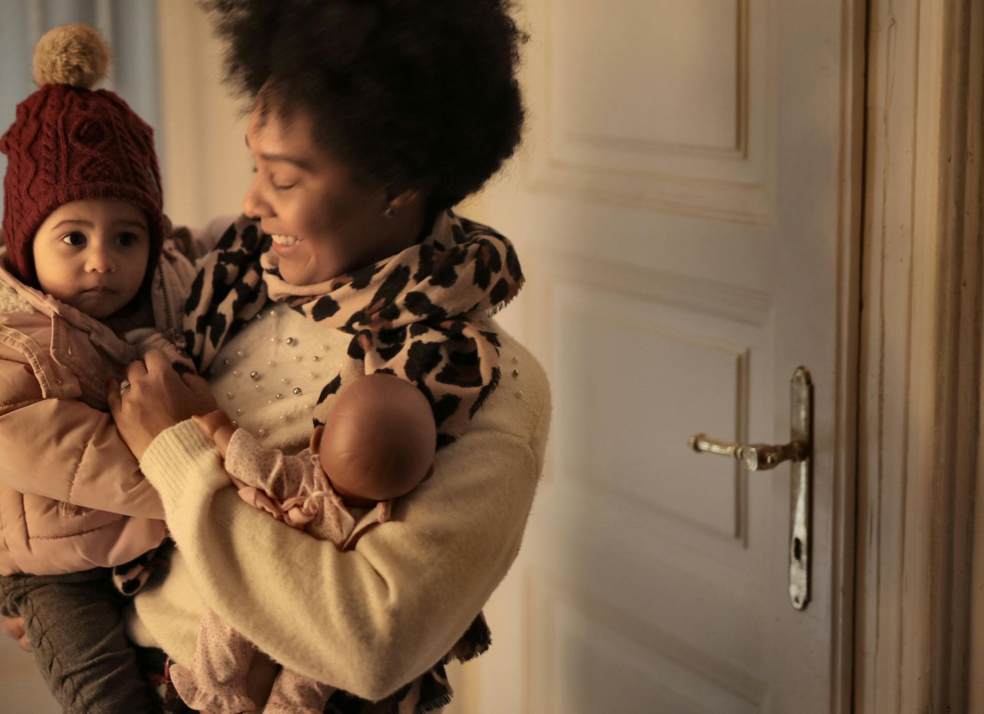 Cheerful ethnic woman hugging little kid while standing in room with toy and looking at clothes of baby while preparing to walk